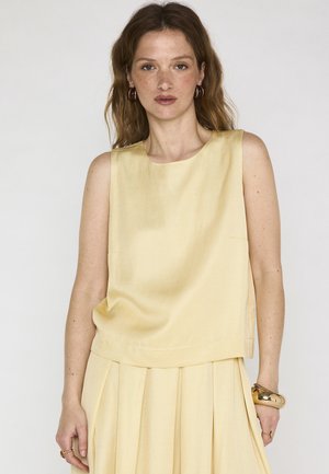 Femme aux cheveux bruns ondulés portant un haut jaune pâle sans manches et une jupe plissée, boucles d'oreilles en or en forme d'anneaux, bracelet en or et bagues, fond neutre.