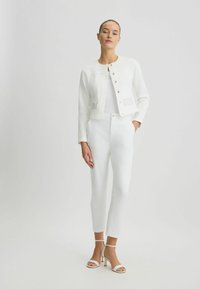 Chaqueta corta de lino blanca con cierre de botones y bolsillos texturizados, combinada con pantalones tailored blancos y tacones altos blancos con tiras.