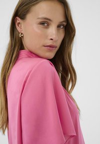 Blusa rosa con un tejido suave y fluido y mangas anchas, que presenta un escote alto y un discreto pliegue en el hombro. Pendientes de aro dorados.