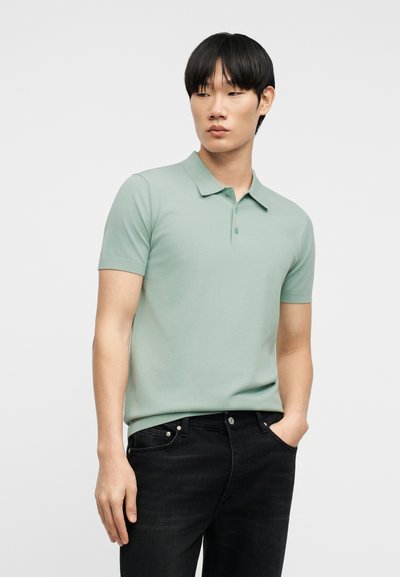 Jeune homme aux cheveux noirs portant un polo vert clair à manches courtes et un jean noir, debout avec une main dans la poche, sur fond blanc.