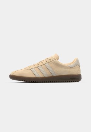 Sneaker en suede beige avec trois bandes argentées, semelle plate en caoutchouc marron, bout arrondi et design minimaliste.