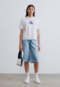 Λευκό βαμβακερό T-shirt με μπλε λογότυπο, συνδυασμένο με ανοιχτόχρωμα τζιν κολότσε. Συμπληρωμένο με ασημένια τσάντα και λευκά αθλητικά παπούτσια.