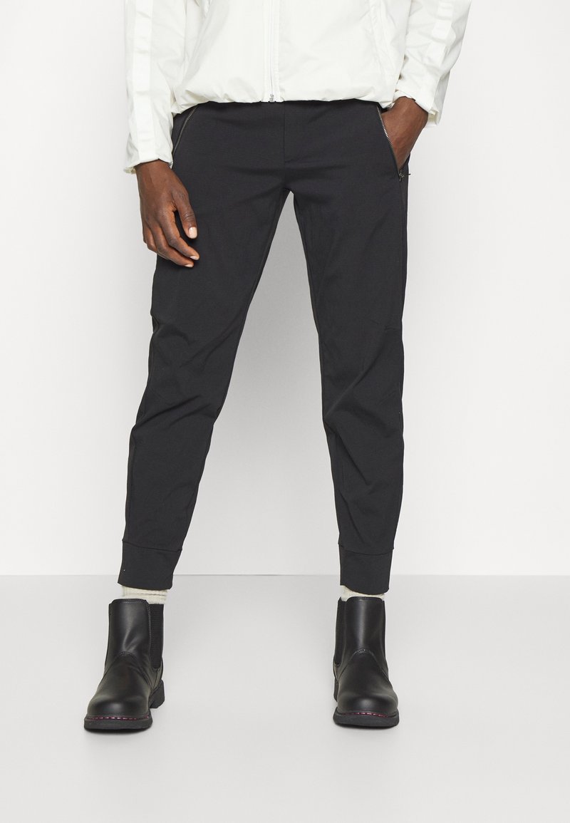 Personne portant un pantalon noir fuselé avec des poches latérales zippées, des chaussettes blanches, des bottines noires et une veste blanche, les mains dans les poches.