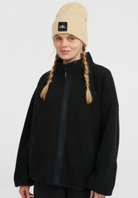Black fleece jacket with a high collar and full zip. Features side pockets, dropped shoulders, and a textured surface. Beige knit beanie.
