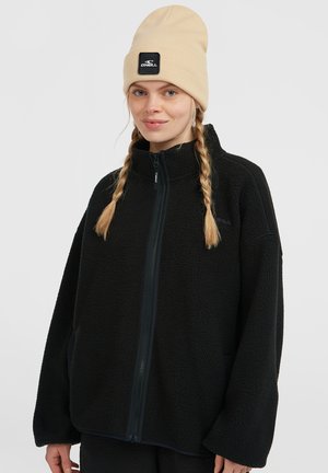 Black fleece jacket with a high collar and full zip. Features side pockets, dropped shoulders, and a textured surface. Beige knit beanie.