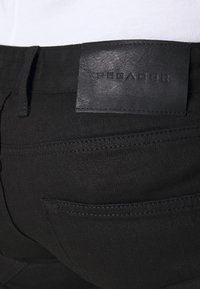 Närbild av bakfickan på svarta byxor med sydda detaljer och en svart lädermärke som säger "PEGADOR."