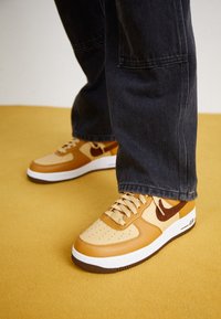 Baskets Nike Air Force 1 en cuir tan et beige avec un bout perforé, accents marron foncé, semelle intermédiaire blanche et semelle extérieure marron contrastante.