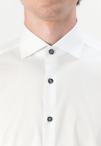 Chemise blanche à col classique, avec des boutons noirs et un tissu lisse et texturé. Les boutons sont alignés sur le devant.