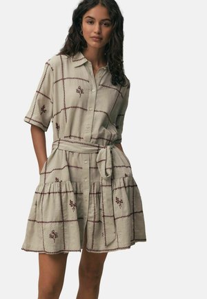 Nainen, joka pukeutuu beigeen lyhythihaiseen napitettuun mekkoon, jossa on ruskeaa kukkaompelua, solmittu vyö vyötäröllä ja kerroksittainen helma, seisoo käsien ollessa taskuissa.