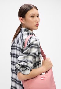 Bolso de hombro rosa hecho de material suave, con una textura lisa y un parche con logo; llevado sobre una camisa de cuadros en blanco y negro.