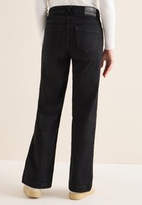 Jeans negros de pierna ancha con cintura ajustable, que cuentan con dos bolsillos traseros y una textura suave. El modelo lleva zapatillas beige.