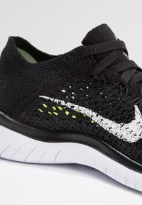 Zwarte gebreide sportschoen met witte swoosh-logo, groene accenten en een gestructureerde afwerking. Heeft een flexibele witte zool met groeven voor grip.