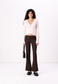 Helles rosa Spitzen-Wickeloberteil mit langen Ärmeln, kombiniert mit braunen Schlaghosen mit einer Doppelknopfverschlüssen. Die Accessoires umfassen eine schwarze Handtasche.