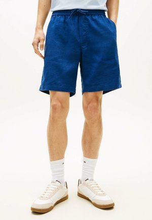 Mann trägt blaue Shorts mit Kordelzug, weiße Sportsocken und weiße Turnschuhe, steht vor einem schlichten hellen Hintergrund.