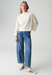 Witte sweater met een ronde hals en doorlopende schouders, gecombineerd met lichtblauwe wijde jeans en witte sneakers met zwarte strepen.