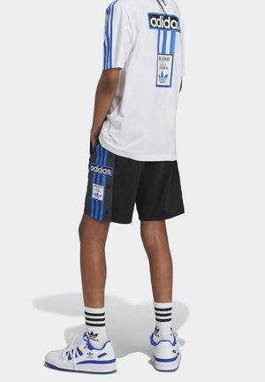 Person wearing white and black Adidas sportswear with blue stripes, standing with hands in pockets, wearing white Adidas sneakers and striped socks.