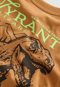 Braunes Baumwoll-T-Shirt mit einem schwarzen Dinosaurier-Design und grünem Text, auf dem "Tyrannenechse" und "68 Millionen Jahre" prominent dargestellt sind.