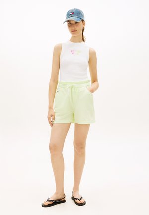 Giovane donna con indosso un top bianco senza maniche, pantaloncini verdi chiaro con coulisse, sandali neri e un cappellino di jeans blu, in piedi con una mano in tasca.