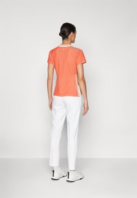 Πορτοκαλί κοντομάνικο μπλουζάκι με στρογγυλή λαιμόκοψη, με υφή και λεπτομέρεια λογότυπου στο πλάι. Συνδυάζεται με λευκά παντελόνια και ανοιχτόχρωμα υποδήματα.