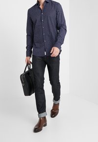 Camicia blu navy con motivo geometrico bianco, con bottoni, abbinata a jeans scuri arrotolati ai polsi, scarpe in pelle marrone e una borsa nera.