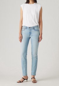 Mujer vestida con una blusa sin mangas blanca, jeans ajustados de color azul claro y sandalias negras de tacón bajo con tiras, de pie contra un fondo liso.