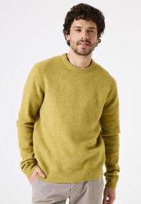 Homme aux cheveux bouclés portant un pull à col rond jaune moutarde et un pantalon gris clair, se tenant contre un fond blanc uni.
