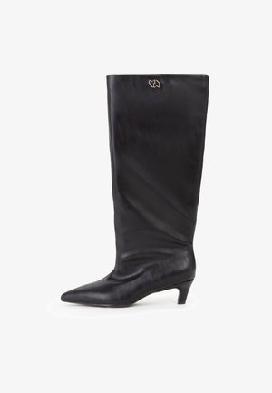 Bota negra de cuero hasta la rodilla con punta afilada y un pequeño tacón cónico. Presenta un acento con el logo dorado en la parte superior. Textura suave.