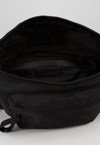 Bolsa de tela negra con un exterior texturizado. La cremallera abierta revela un interior liso y una etiqueta con la inscripción "ZAIGN". Diseño minimalista.