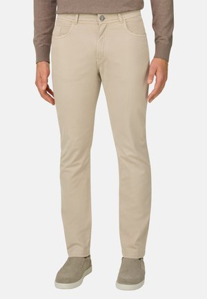 Man draagt beige, nauwsluitende broek, taupe lange mouwen shirt en grijze instapschoenen, staand tegen een effen achtergrond.