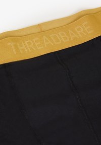 Schwarze Unterwäsche mit einem gelben elastischen Bund, auf dem der eingeprägte Text "THREADBARE" zu sehen ist. Auffällige Nahtdetails an den Seiten.