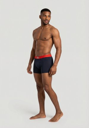 Atletyczny mężczyzna stojący boso, ubrany w granatowe bokserki z czerwoną gumką, patrzący w bok na jednolitym tle.