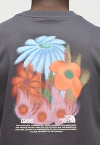 Πίσω μέρος γκρι πουκαμίσου με πολύχρωμα αφηρημένα λουλούδια, "1966" και το λογότυπο της The North Face με κείμενο αποστολής.