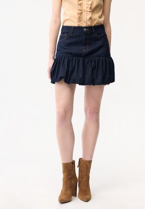 Kvinde iført en mørk denim mini nederdel med flæser, tan ærmeløs bluse og brune ruskind ankelstøvler på hvid baggrund.