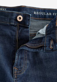 Jean denim bleu foncé avec une coupe régulière. Il possède une fermeture à glissière, un bouton en cuivre et des coutures contrastantes orange. Doublure intérieure visible.