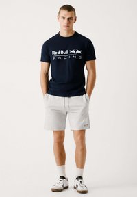 Tamsiai mėlynas marškinėliai su apvaliu kaklu ir "Red Bull Racing" logotipu, derinami su šviesiai pilkomis šortais ir baltais sportiniais batais su juodomis detalėmis.