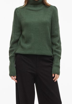 Kvinde iført en grøn strikket rullekravesweater med lange ærmer og sorte løstsiddende bukser mod en hvid baggrund.