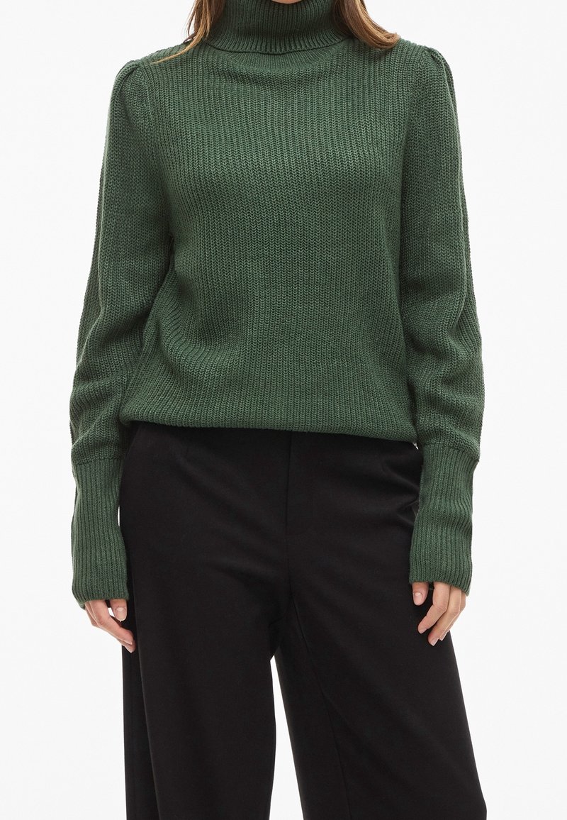 Femme portant un pull en maille vert à col roulé avec des manches longues et un pantalon noir ample sur fond blanc.
