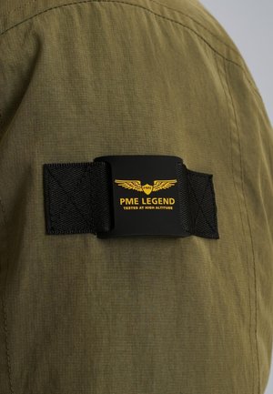 Un tissu vert à finition texturée présente une bande noire avec un patch étiqueté "PME LEGEND" en texte jaune et logo ailé.