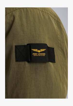 Tessuto verde con finitura testurizzata presenta un'etichetta a fascia nera con la scritta "PME LEGEND" in giallo e un logo con ali.