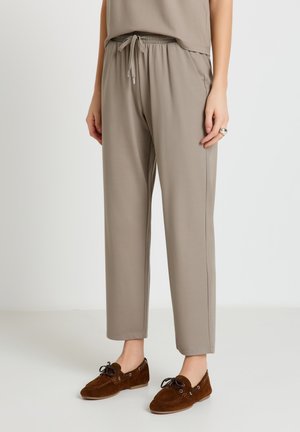 Pantaloni beige con coulisse indossati con top abbinato e mocassini stringati in camoscio marrone su sfondo chiaro.