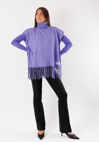 Poncho viola con collo a dolce vita e dettaglio con frange sull'orlo e fessure laterali, indossato sopra una maglietta a maniche lunghe, abbinato a pantaloni neri a zampa.