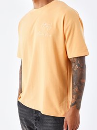 Korte mouw, perzikkleurig T-shirt van katoen; heeft een wit hartontwerp met een wereldkaart en tekst op de borst.