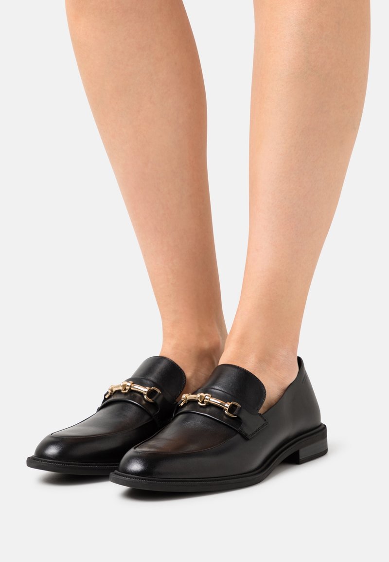 Mocassins en cuir noir avec détail de chaîne dorée portés pieds nus, montrés en vue de face sur un fond clair uni.