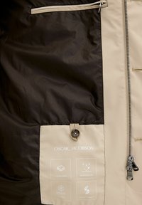 Giubbotto idrorepellente con tessuto a due strati, in marrone e beige. Include una cerniera, dettagli con bottoni e etichette con specifiche del tessuto.