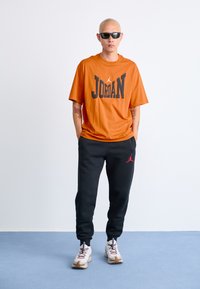 Mand med barberet hoved iført sorte solbriller, orange Jordan T-shirt, sorte joggebukser med rødt logo og hvide sneakers på lys blå gulv.