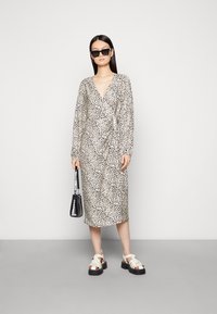 Vestido envolvente de mangas longas em estampado de leopardo, com decote em V profundo, até ao joelho, combinado com sandálias de plataforma e uma pequena bolsa preta.