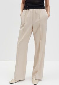 Pantaloni beige a gamba larga con pieghe frontali indossati da una persona in top nero, con le mani in tasca, abbinati a scarpe a punta color crema.