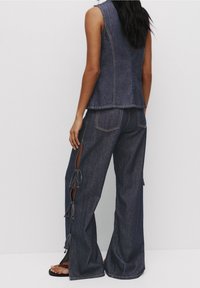 Ensemble en denim comprenant un gilet et un pantalon à jambes larges, de couleur bleu foncé. Le gilet présente un design court, tandis que le pantalon possède des fentes latérales avec des liens et des coutures visibles.