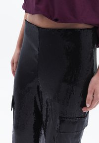 Zwarte paillettenrok met een gestructureerd oppervlak, voorzien van een zijzak en een getailleerde silhouette, gecombineerd met een losse paarse top.