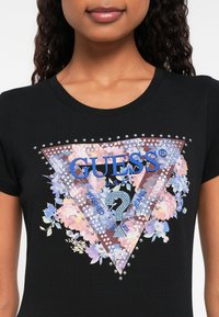 Ung kvinne iført svart t-skjorte med blomstrete og naglede trekant Guess-logo på brystet.